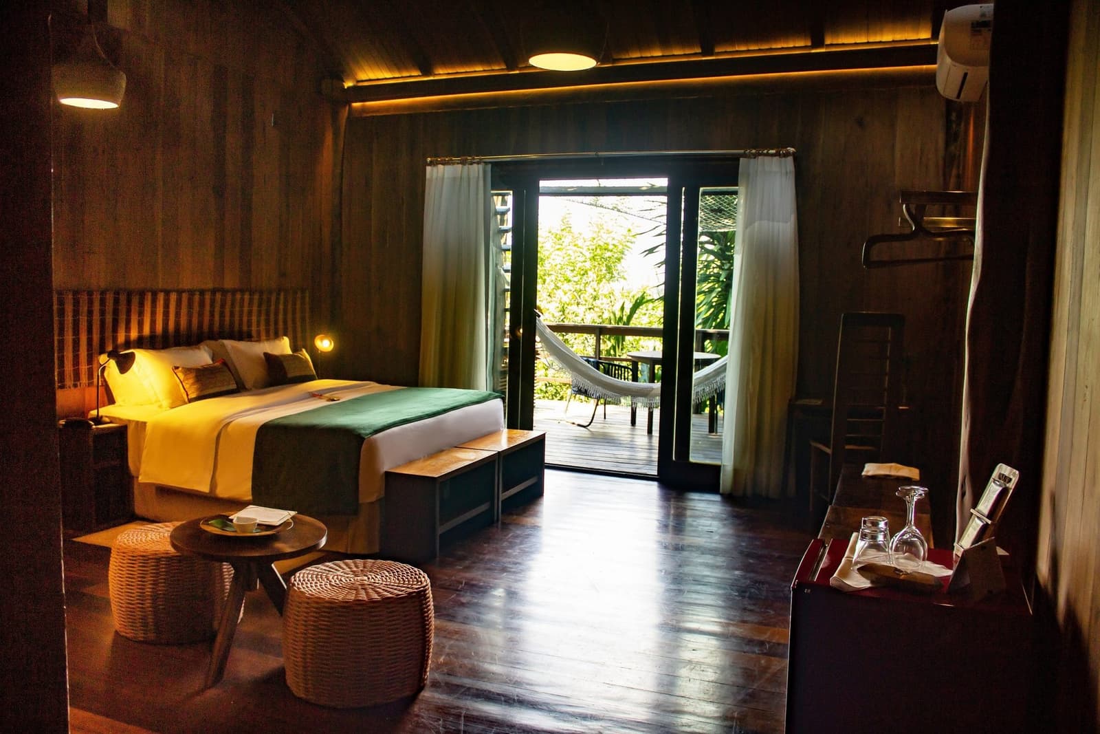 A hammock on the balcony: the Amazon's way of saying slow down. Warmly lit luxury amazon lodge brazil suite with a double bed, wicker ottomans, and open French doors leading to a private hammock balcony surrounded by tropical greenery