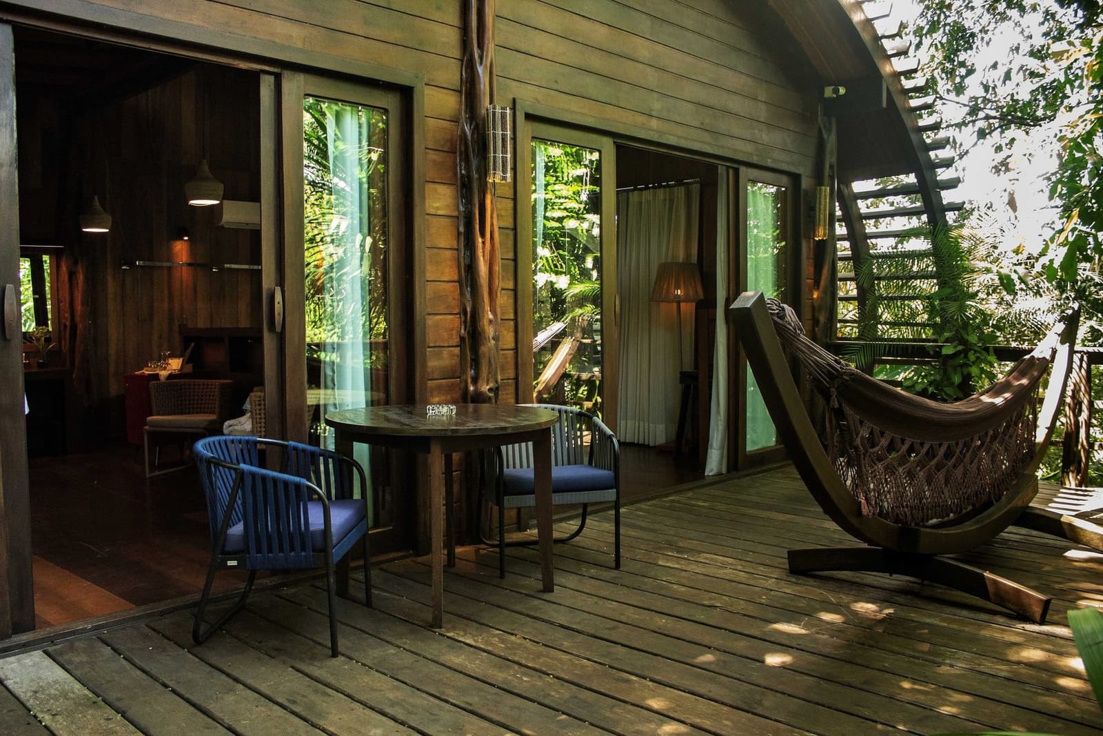 The hammock is right there. The forest does the rest. A private wooden deck at a luxury amazon lodge brazil featuring a round table with two chairs, a freestanding hammock, and sliding glass doors opening to a forest-surrounded cabin