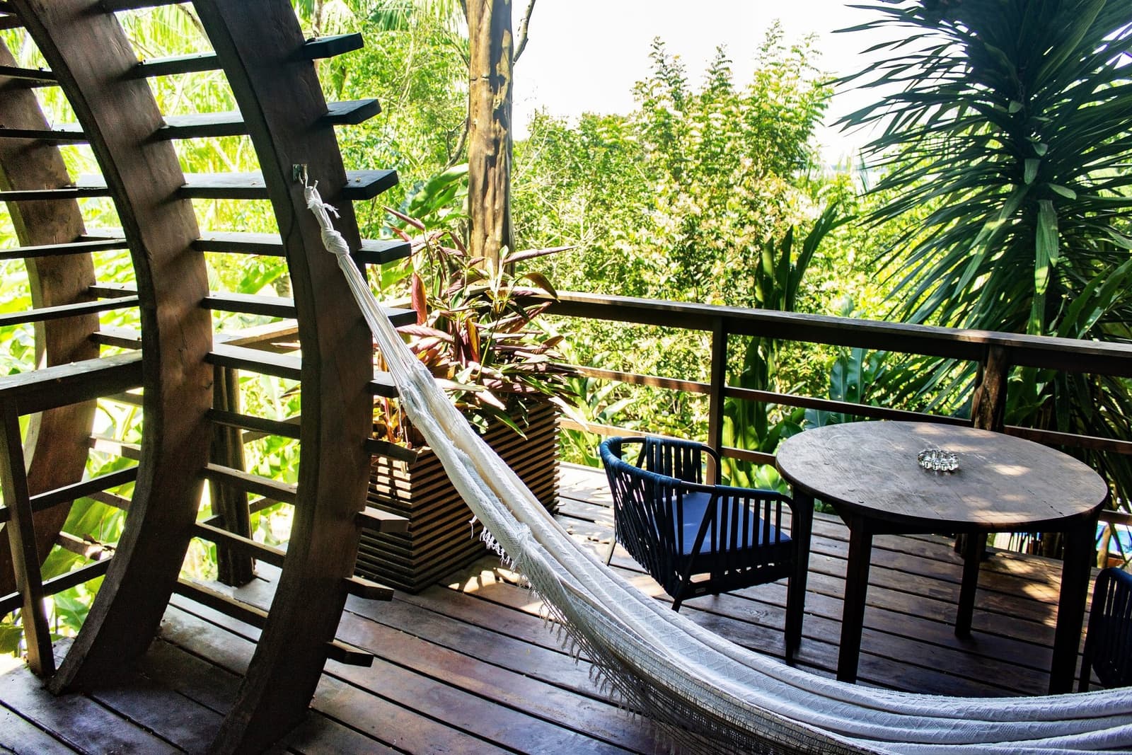 Your afternoon plans, sorted: hammock first, everything else later. A white fringed hammock and round hardwood table on a curved timber deck at a luxury amazon lodge brazil, surrounded by lush tropical greenery