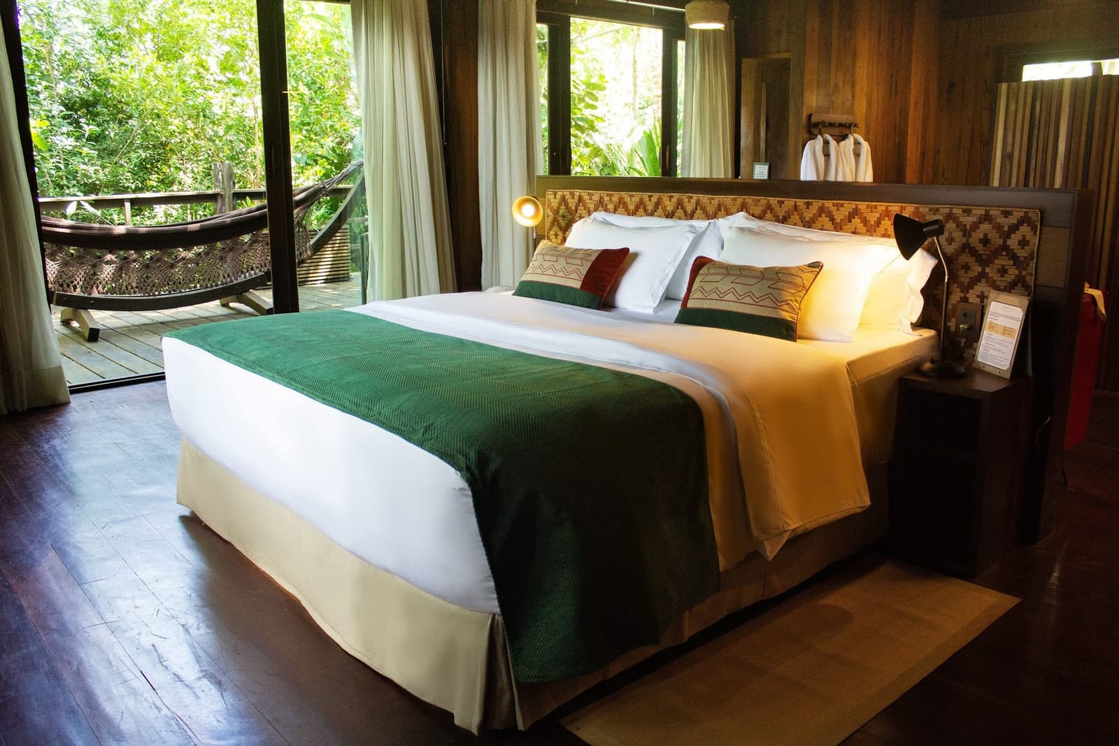Warm wood, forest green, and the sound of the rainforest just beyond the door. King bed with a woven geometric headboard and green velvet runner in a luxury amazon lodge brazil cabin, with a private hammock deck framed by lush rainforest visible through floor-to-ceiling glass doors