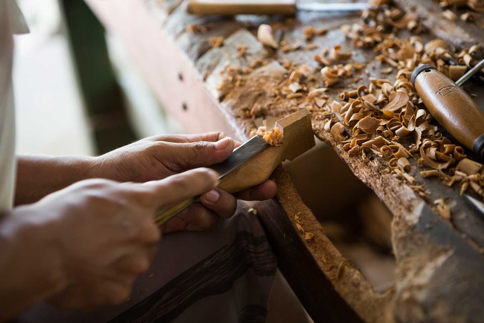 Slow craft, sharp tools, and a workbench that tells the whole story. Close-up of hands using a chisel to carve a wooden piece at a workbench covered in fresh wood shavings, reflecting the local artisan craft traditions near a luxury Amazon lodge in Brazil
