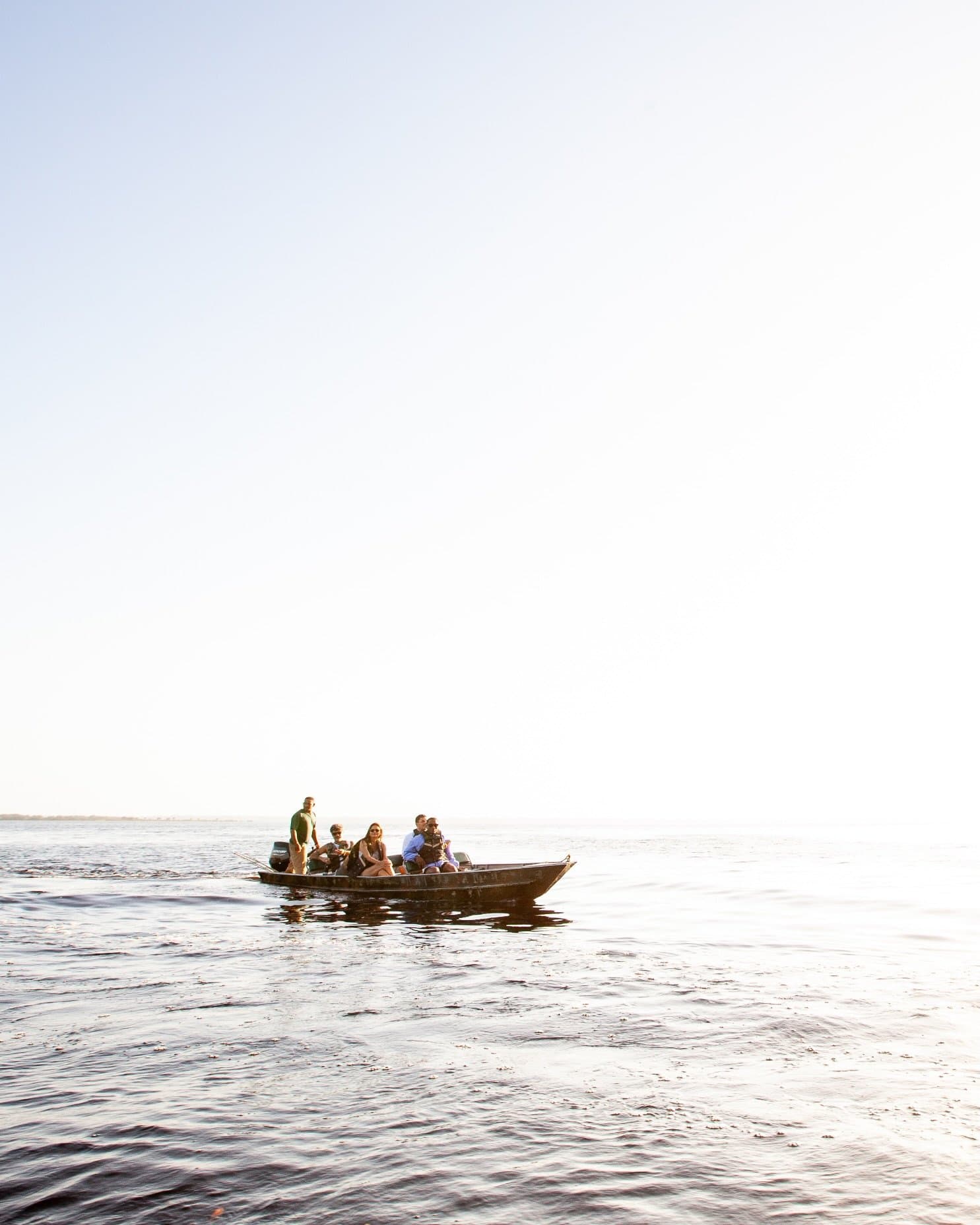 Out here, the river is the road and the horizon keeps moving. A small wooden motorboat carrying a group of travellers glides across a vast, calm expanse of dark river water under a bright hazy sky on a luxury Amazon lodge Brazil excursion