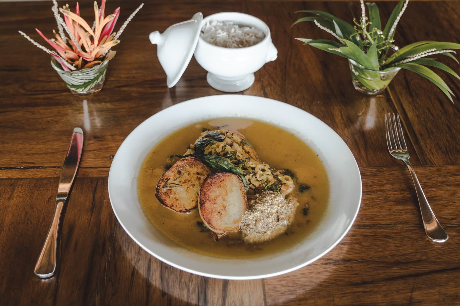 Regional cooking this good makes you forget there's a whole rainforest outside. A white bowl of golden broth with seared medallions, greens, and manioc flour served at a luxury Amazon lodge Brazil restaurant, with a lidded rice tureen and tropical plant arrangements on a polished hardwood table