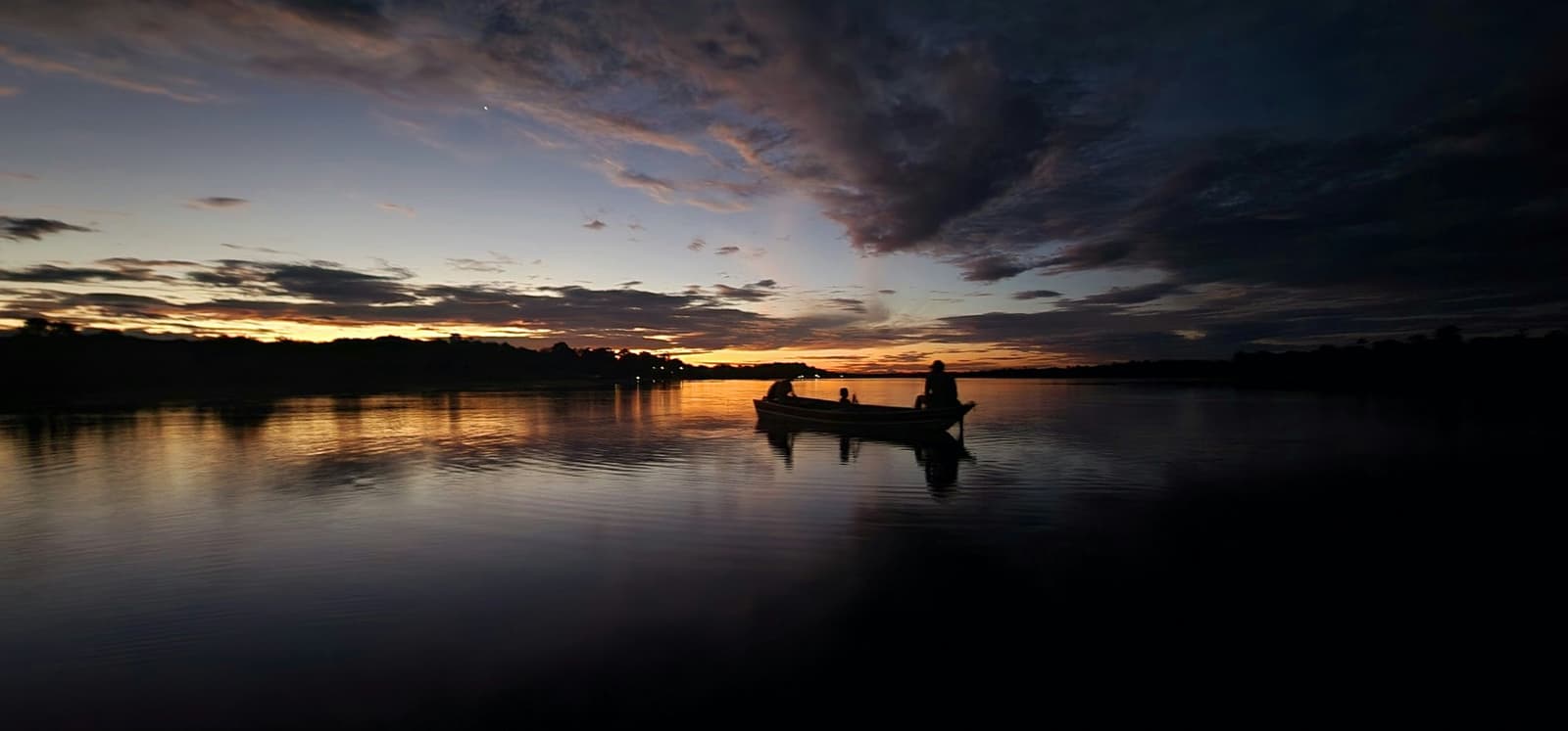 The river holds the whole sky, and the boat knows exactly where it's going. Silhouette of three people in a small wooden boat on a glassy river at dusk, golden and violet clouds reflected on the still water near a luxury Amazon lodge Brazil experience