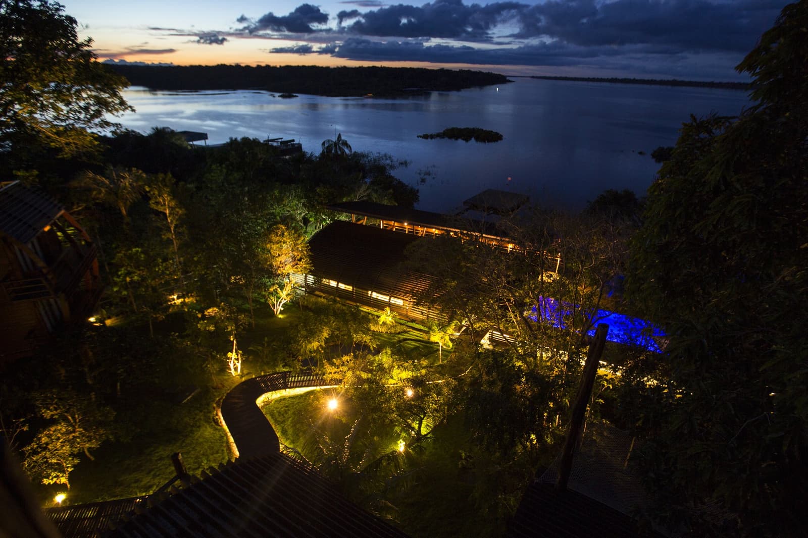 Sunset over the river, and the lodge lights up like it's been waiting for you. Aerial twilight view of a luxury Amazon lodge Brazil, with warm golden lights illuminating timber buildings, a winding boardwalk and lush jungle garden leading down to a wide dark river dotted with forested islands under a dramatic dusk sky