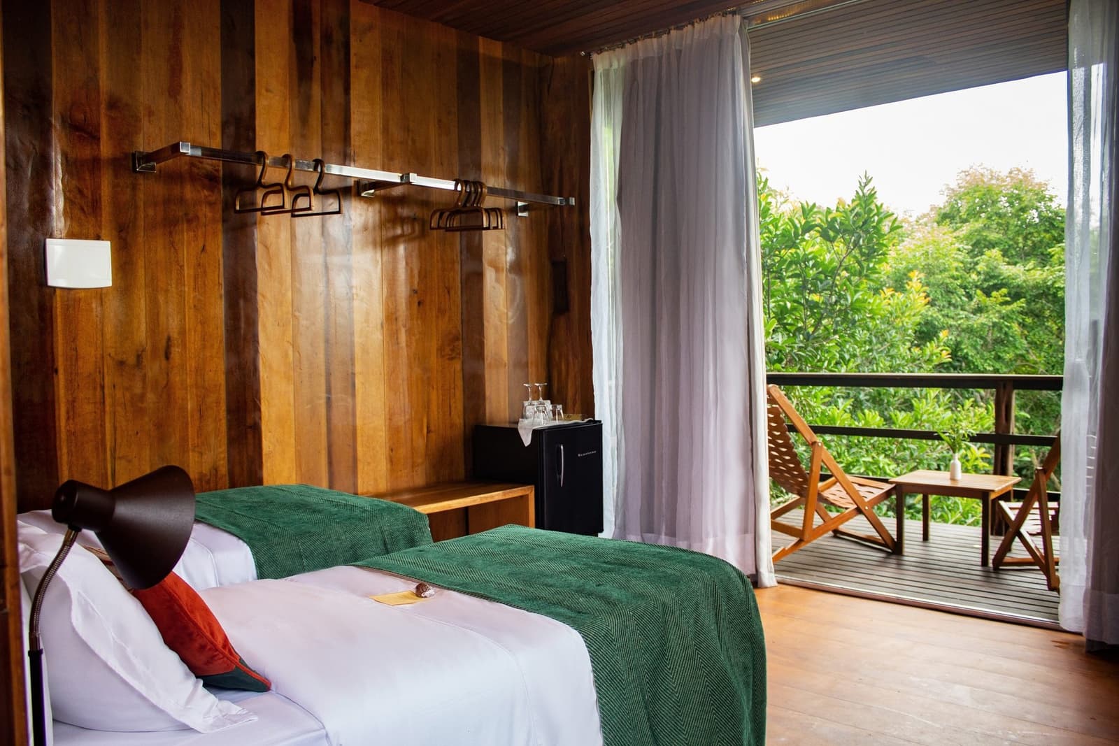 Your morning view is all green canopy, before you've even had coffee. Twin beds with green throws in a wood-panelled room at a luxury amazon lodge brazil, opening onto a private balcony with reclining chairs set among lush green treetops