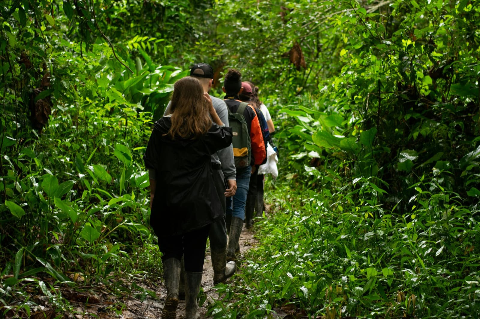 Mud on your boots means you went somewhere real. A small group of travelers in rubber boots walking single-file along a muddy trail through dense green rainforest vegetation on a Manaus jungle tour