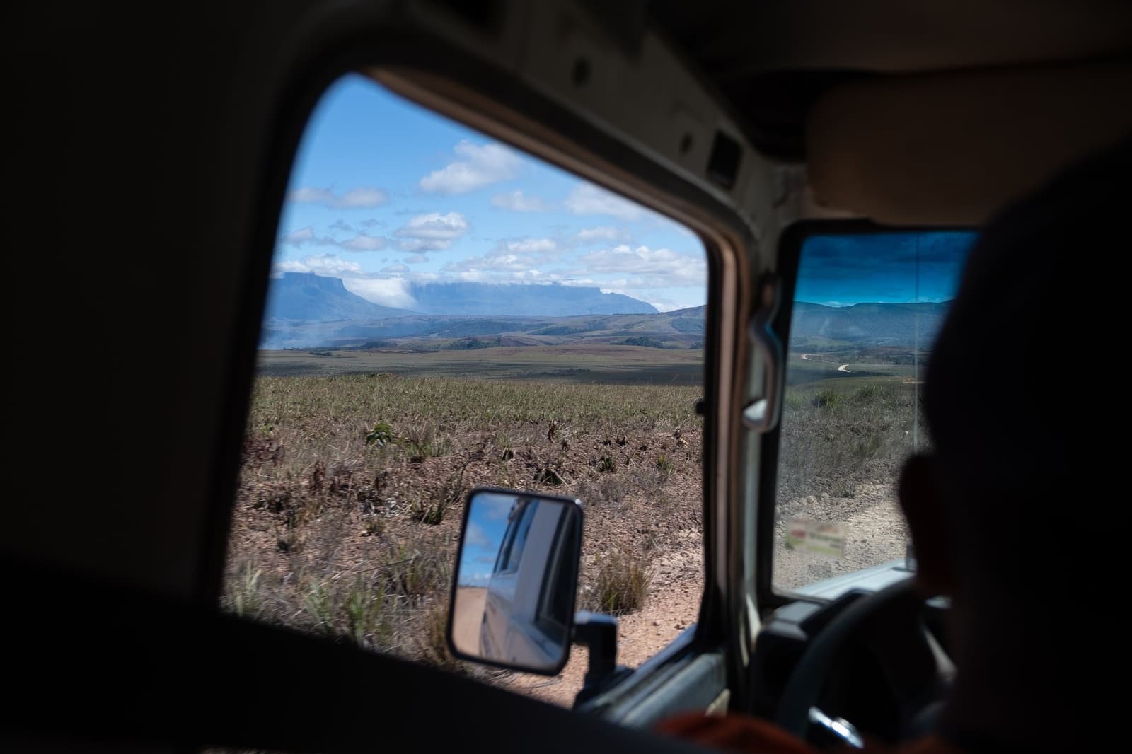 The tepuis appear on the horizon before you've even left the vehicle. Good things take a dirt road. View through a 4x4 vehicle window of the vast Gran Sabana savanna with flat-topped tepui mountains on the horizon, framed by the wing mirror and a dusty dirt track — the approach to the Mount Roraima hike