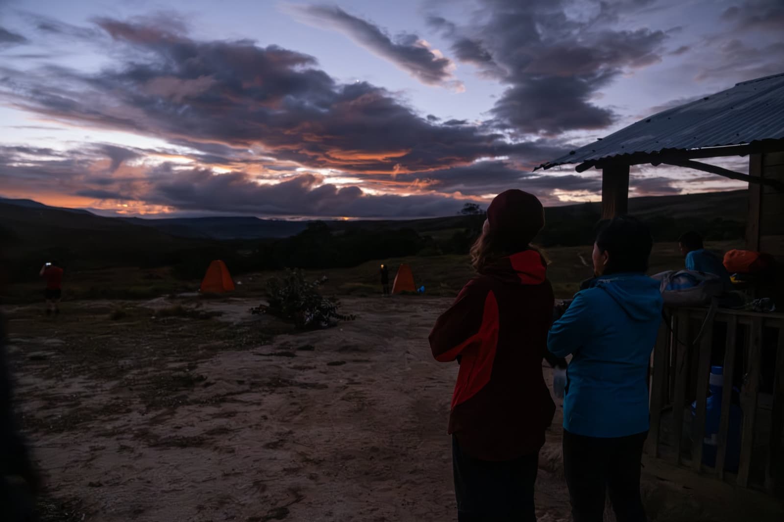 That moment when the sky does all the talking and nobody reaches for their phone. Trekkers in warm jackets watch a vivid orange and purple twilight sky from a remote campsite on the Mount Roraima hike, with orange dome tents glowing in the background