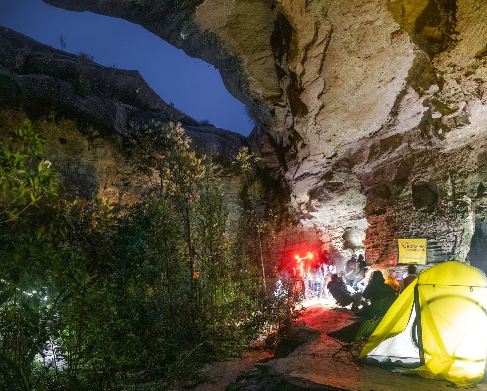 Not every roof needs four walls. This one is 70 million years old. Trekkers camped under a massive sandstone rock overhang at night during the Mount Roraima hike, a glowing yellow tent lit from within beside a group gathered around red lights beneath ancient tepui walls