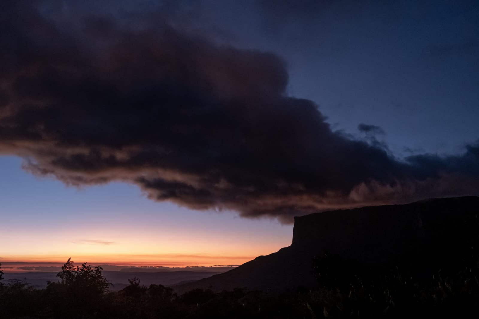 The mountain has its own weather, and it rarely asks permission. A massive dark storm cloud spreads over the silhouette of a flat-topped tepui during a vivid orange and deep blue sunset on the Mount Roraima hike
