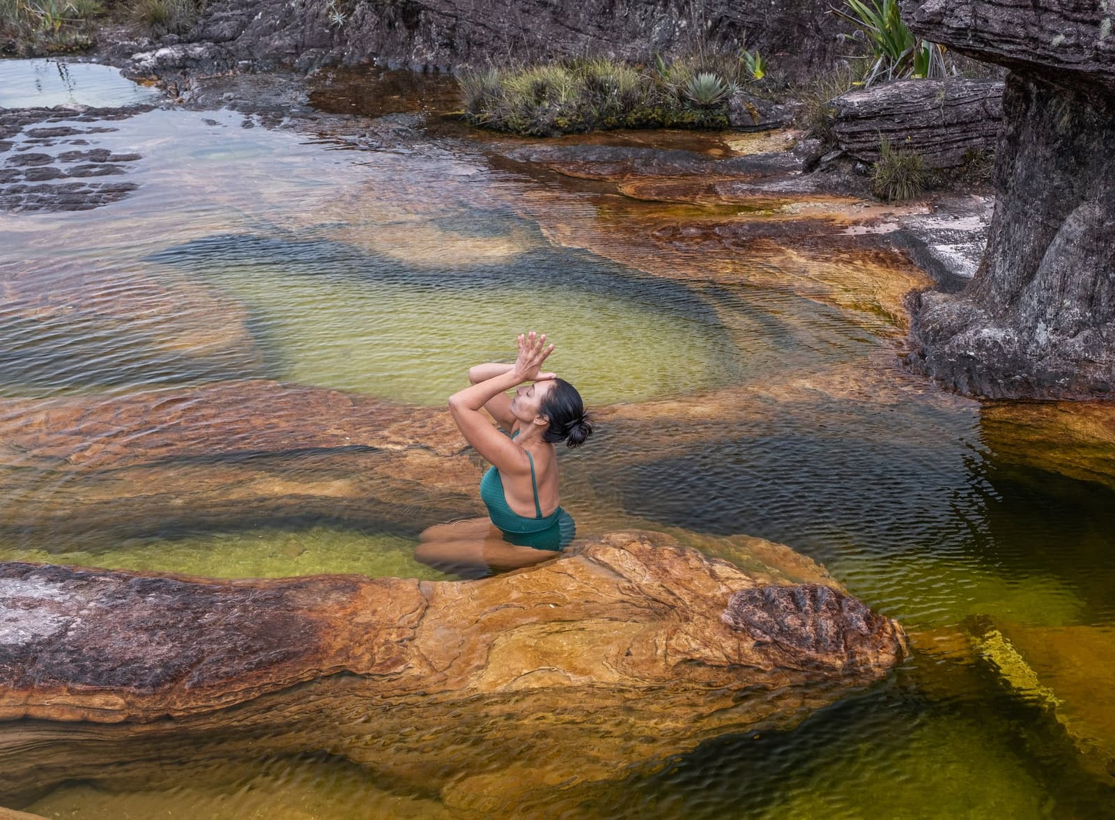 A natural pool billions of years in the making, all to yourself. A woman in a teal swimsuit kneels meditatively in a shallow natural pool on the Mount Roraima hike, surrounded by rust-coloured layered rock and crystal-clear green water