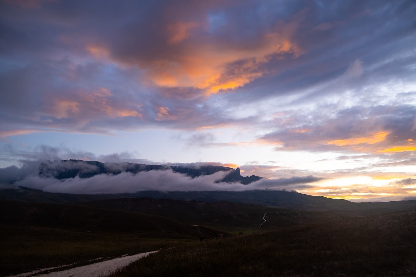 The mountain keeps its secrets under cloud. You earn this light on foot. Dramatic sunset sky with orange and purple clouds billowing above a flat-topped tepui silhouette on the Mount Roraima hike, with a winding dirt track visible across the darkened valley below