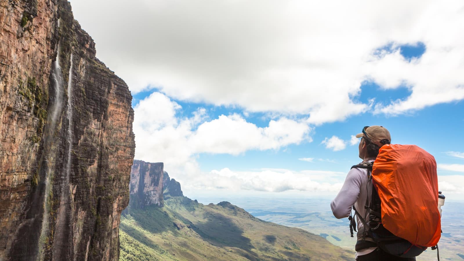 The view earns every blister on the way up. A hiker with an orange-covered backpack stands at the edge of a towering sandstone cliff face on the Mount Roraima trek, gazing across a vast savannah landscape as a thin waterfall cascades down the sheer vertical wall beside them