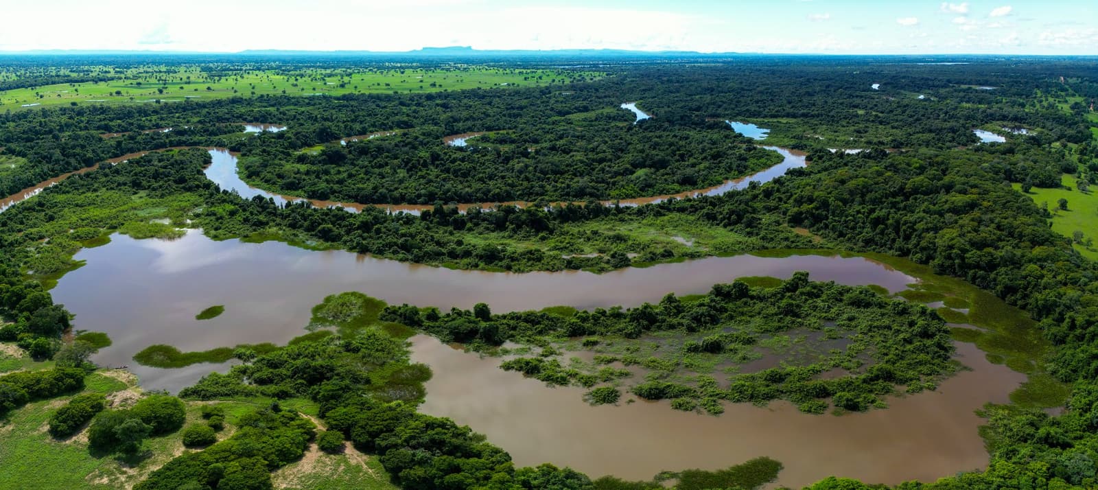 The Pantanal from above: a landscape that thinks in curves, not straight lines. Aerial panorama of a broad meandering river winding through dense tropical forest and flooded plains on a Pantanal Bonito tour, with distant mountains on the horizon