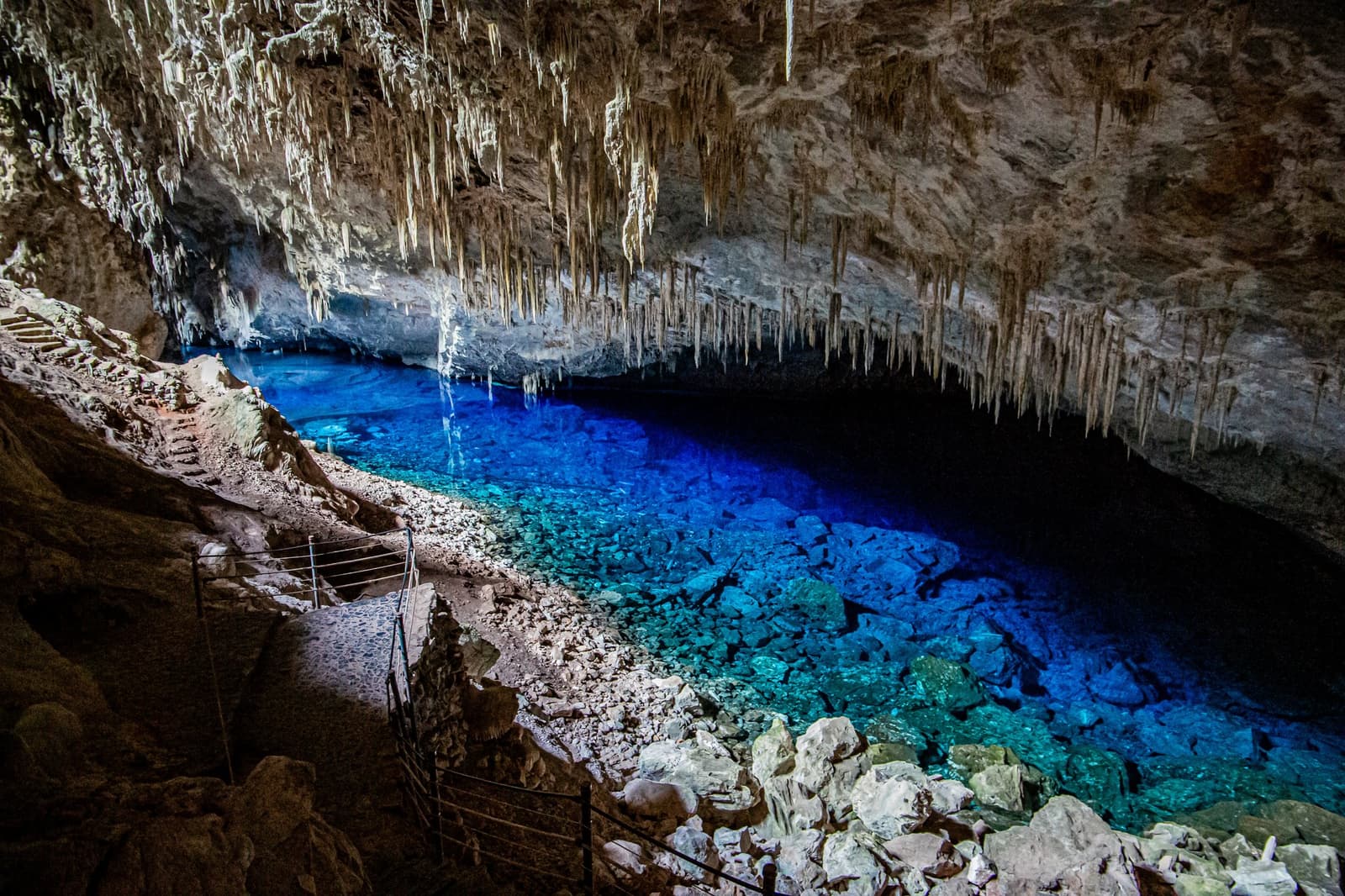 Bonito keeps its best colours underground. A glowing turquoise underground lake inside a limestone cave on a Pantanal Bonito tour, with dense stalactites hanging from the ceiling and a narrow viewing path along the rocky shore