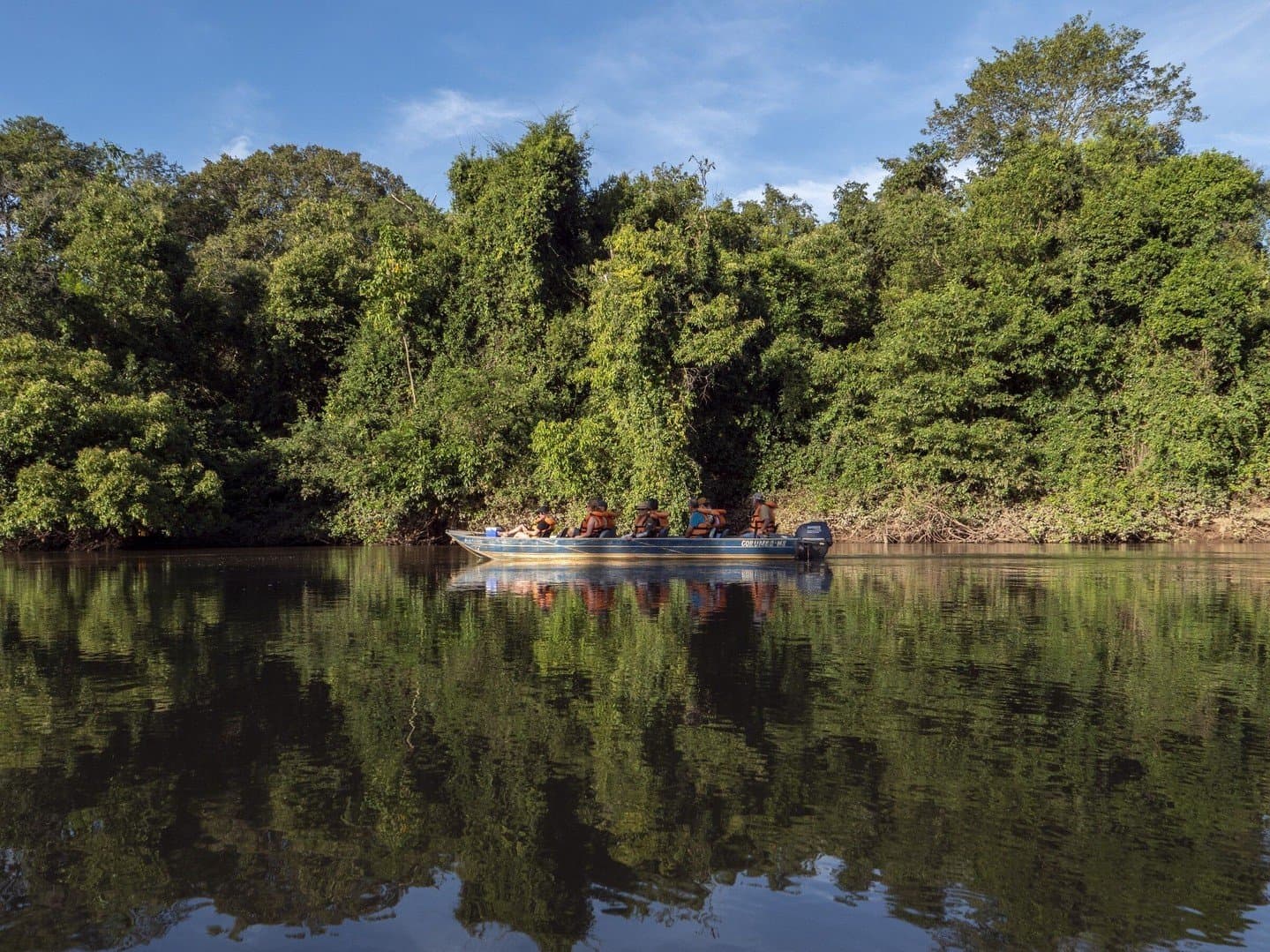 The river knows every secret. Your job is just to listen. A small motorboat carrying wildlife watchers in orange life vests glides along a calm river channel lined with dense green riparian forest on a Pantanal Bonito tour