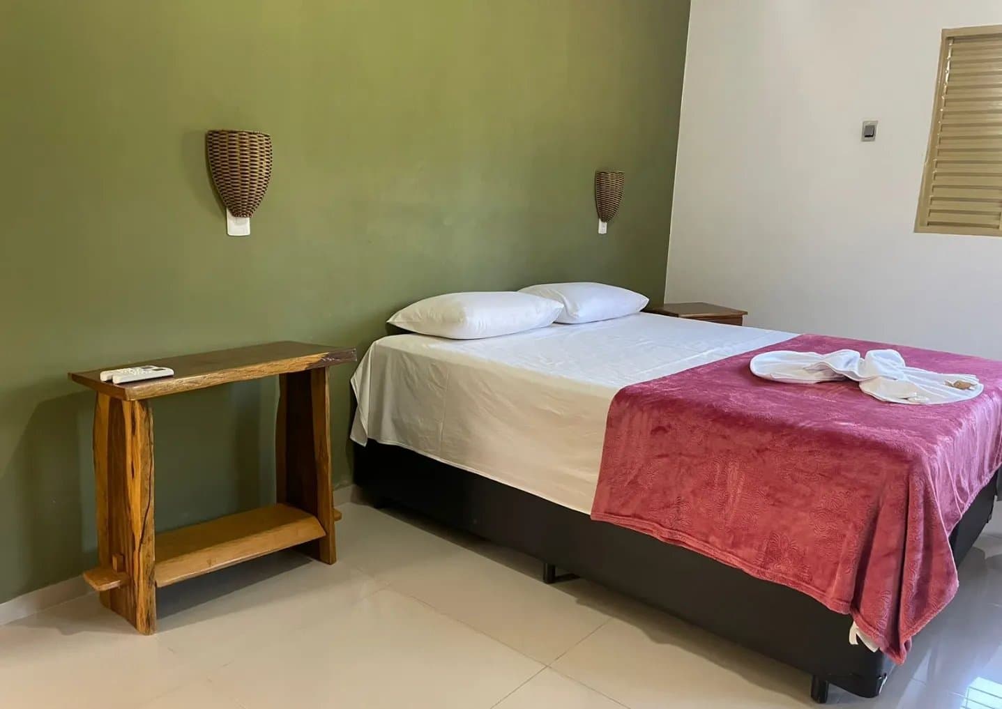Simple, clean, and everything you actually need after a full day in the wild. A clean double room on a pantanal jaguar budget safari, featuring a double bed with white linens and a red throw, wicker wall sconces on a sage-green wall, and a rustic live-edge wooden side table