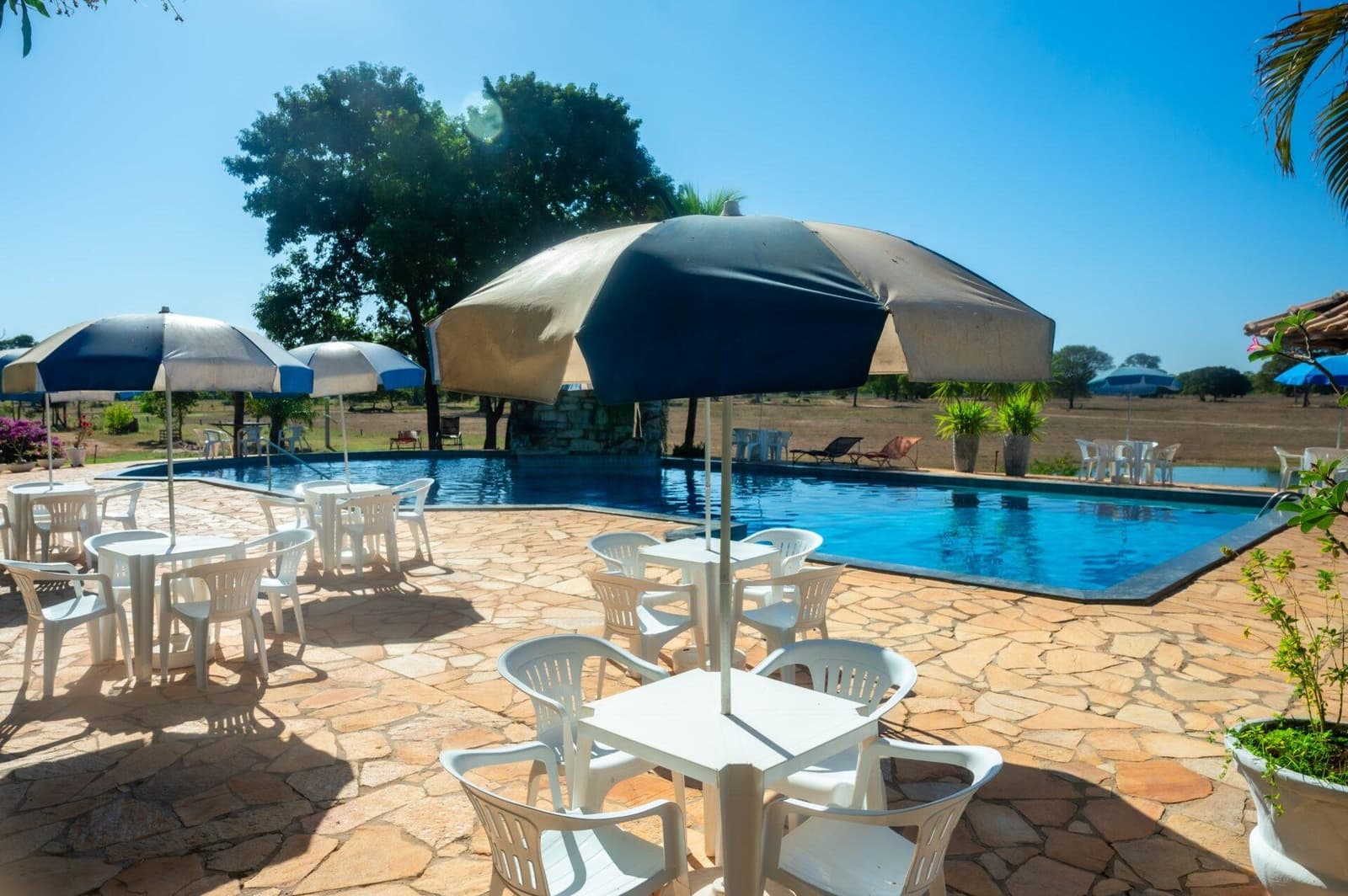 Cold drink in hand, a shady spot by the pool, the afternoon is yours. Sunny outdoor pool terrace with white tables, chairs, and striped umbrellas at a lodge on a pantanal jaguar budget safari