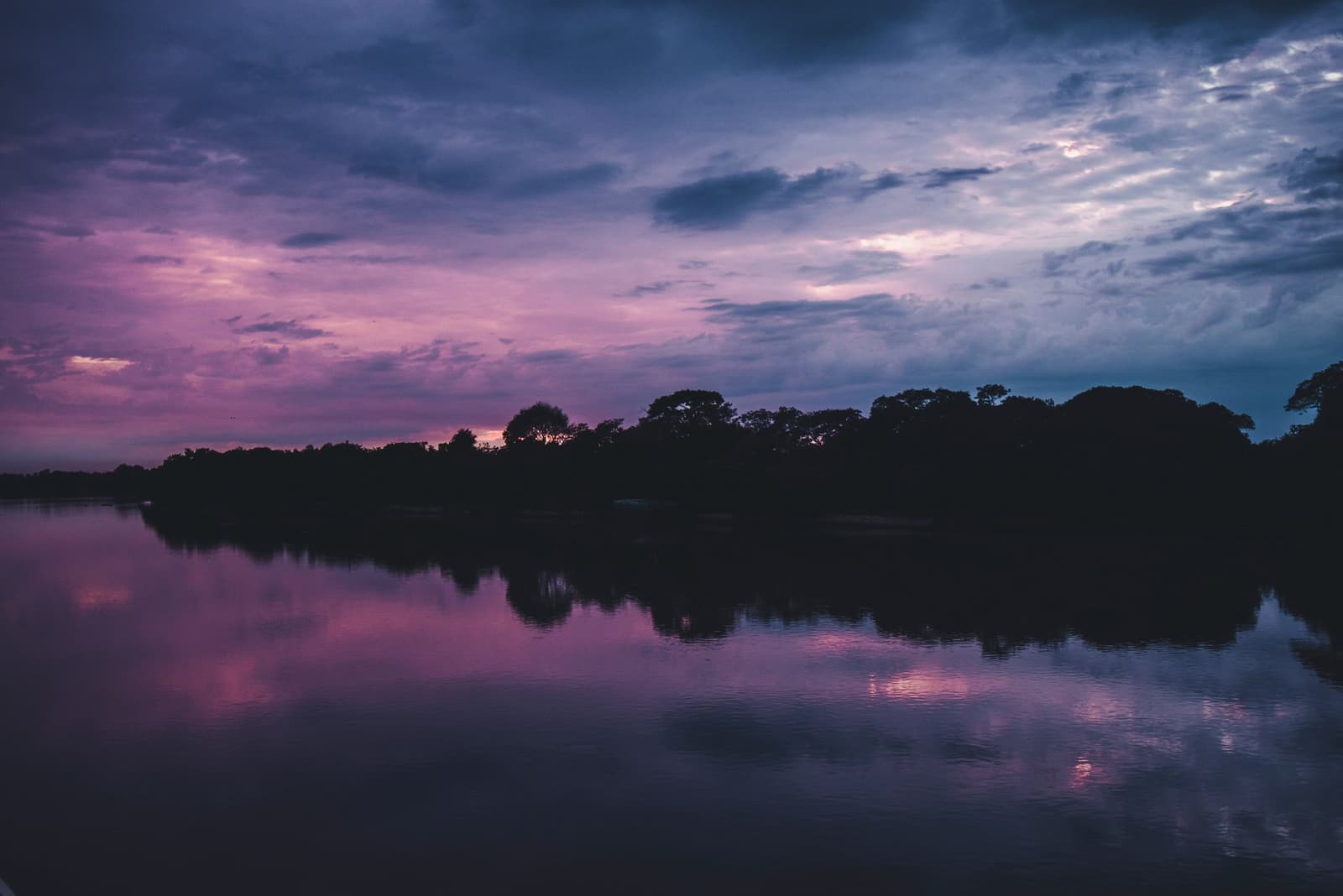 The river holds its breath right after sunset. A vivid purple and pink twilight sky reflected on a still river, with a dark treeline silhouetted along the far bank on a pantanal jaguar budget safari