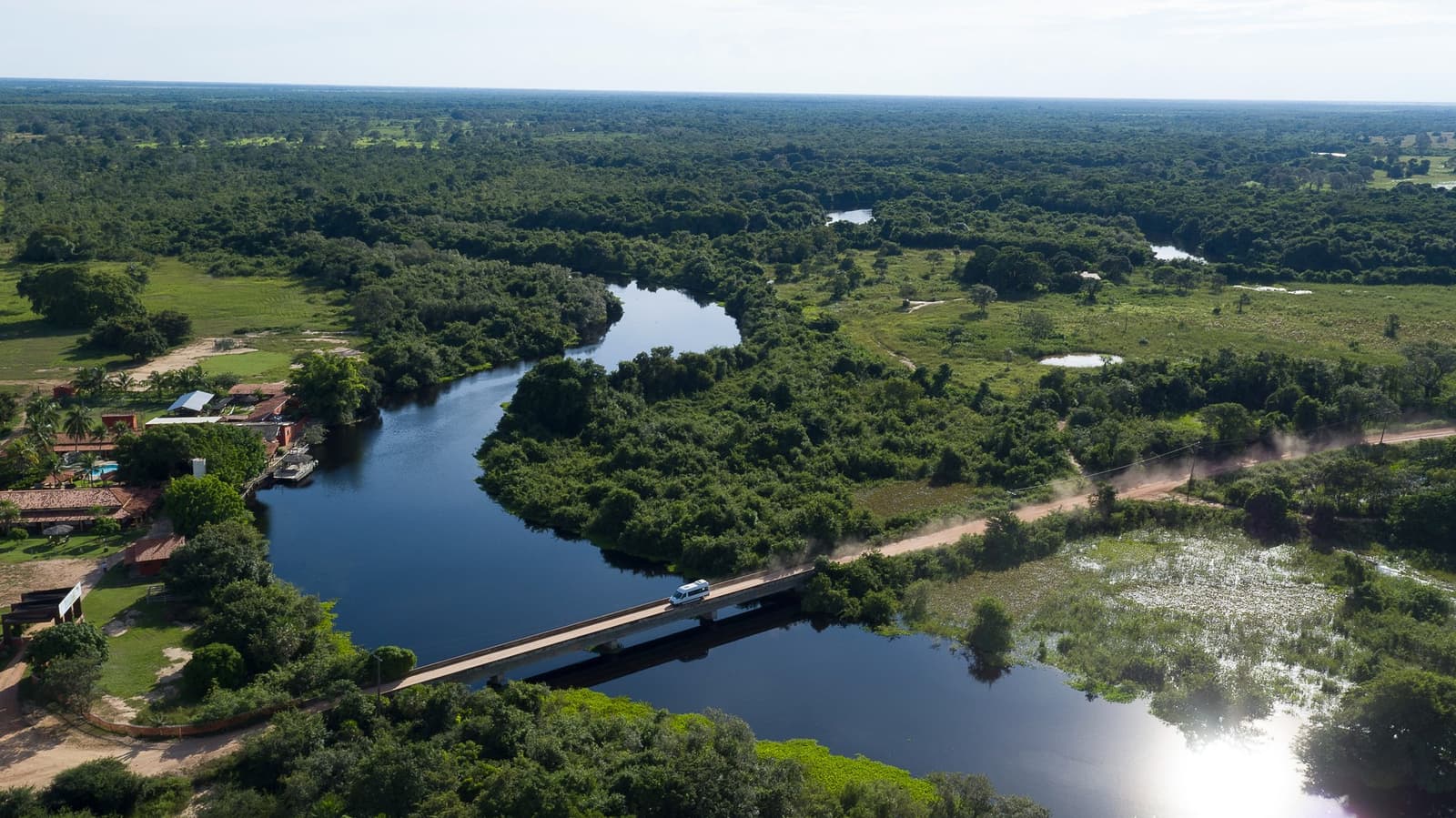 Every Pantanal adventure starts with crossing into a world of its own. Aerial view of a white van crossing a concrete bridge over a winding river surrounded by dense Pantanal wetland vegetation and a rustic riverside lodge, the gateway to a pantanal jaguar budget safari