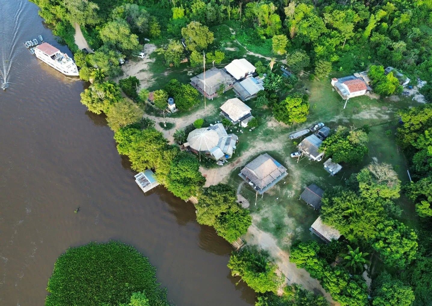 Your base camp sits right where the river decides everything. Aerial drone view of a small riverside lodge tucked into dense green forest along a brown river in the Pantanal, with a multi-deck boat moored at the bank and thatched-roof buildings set among tropical trees
