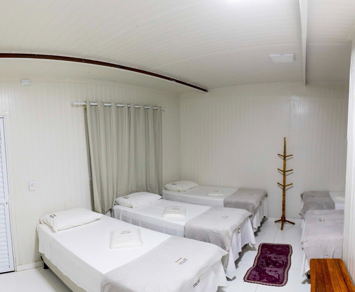 Simple, spotless, and yours after a long day in the field. Clean shared dormitory room with four single beds dressed in white linen, a wooden coat stand, and a curtained window at a Pantanal jaguar budget safari lodge