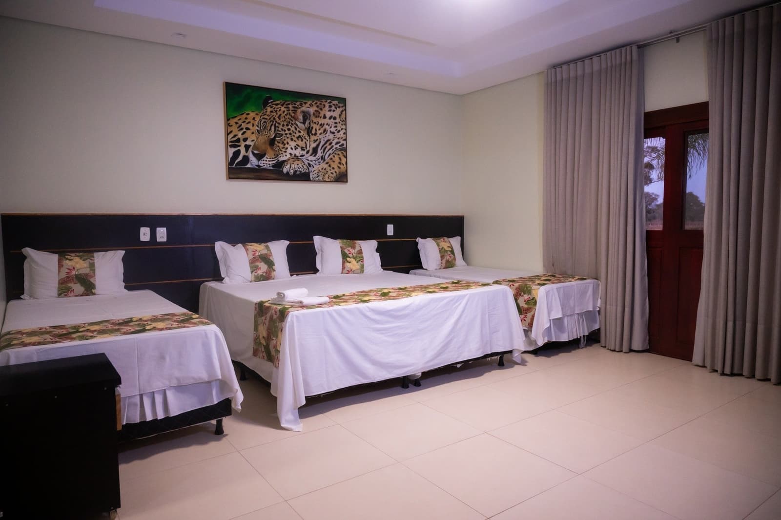 The jaguar watches over you even before the real one shows up. Spacious triple-bed guest room on a pantanal jaguar budget safari, with white linens, tropical-print bed runners, and a jaguar painting above the dark headboard