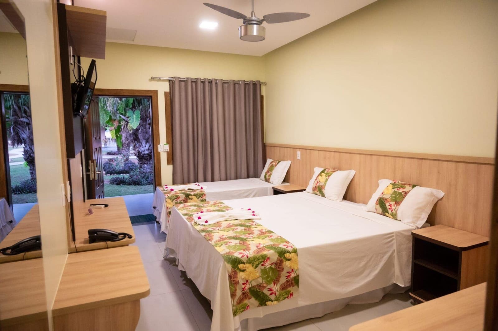 Simple, comfortable, and ready for you after a long day in the field. Triple guest room on a pantanal jaguar budget safari, with two double beds dressed in tropical-print runners, ceiling fan, wall-mounted TV, and an open door revealing lush garden palms beyond