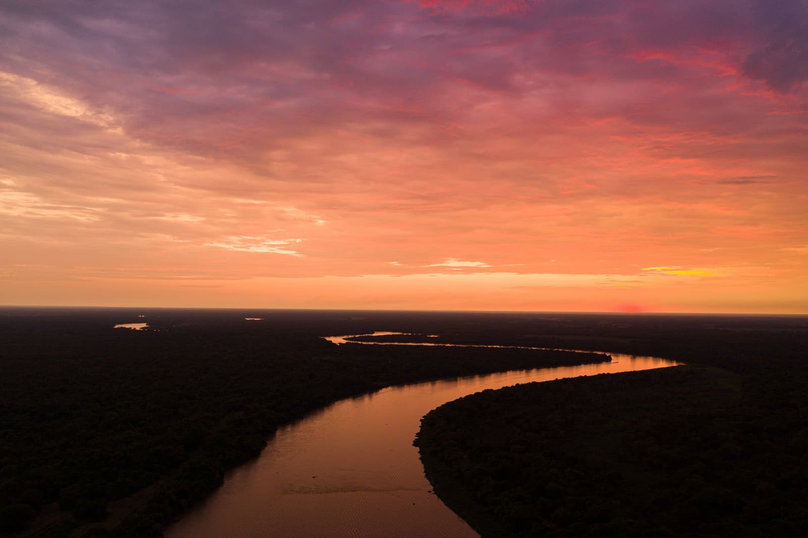 The river knows exactly where it's going, even at dusk. Aerial view of a winding river reflecting a vivid orange and purple sunset sky over vast flat wetlands on a pantanal jaguar safari porto jofre expedition