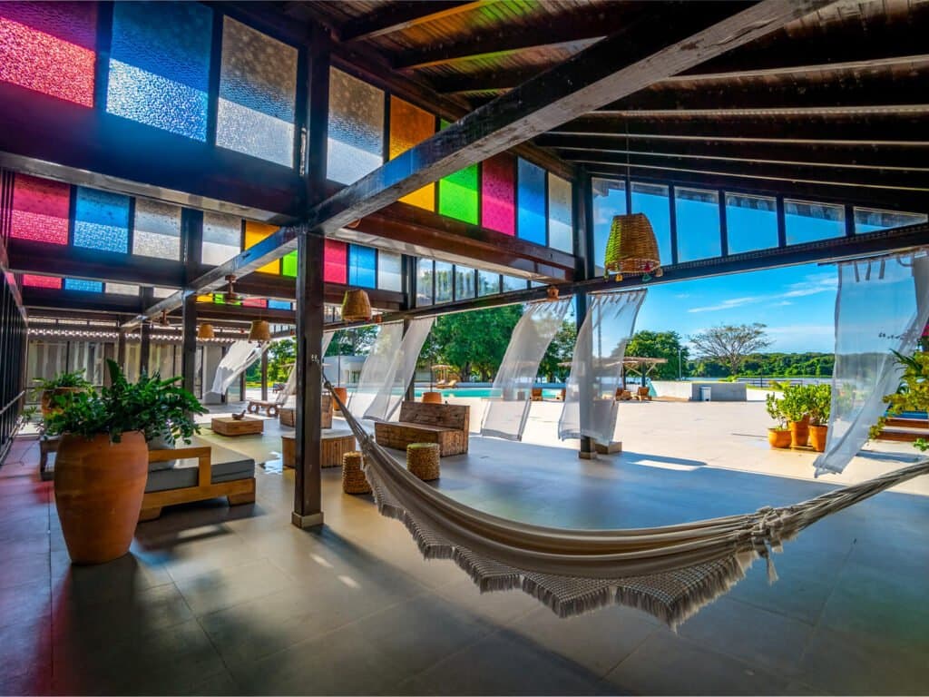 Pick a hammock, ignore the time, repeat. Open-air lodge lounge with a fringed hammock in the foreground, colorful stained-glass panels overhead, rattan pendant lights, and a view across a sunny terrace toward the pool and tree line — a relaxing retreat on a pantanal jaguar safari porto jofre