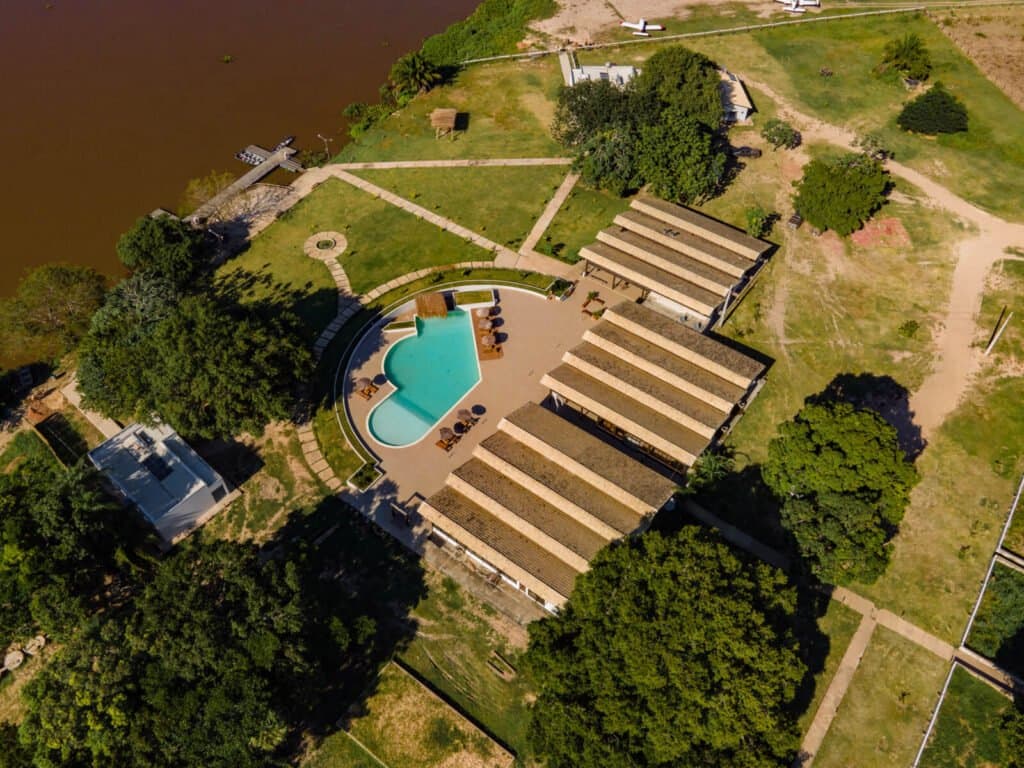 Your base camp, as seen by the birds overhead. Aerial view of a riverfront Pantanal jaguar safari Porto Jofre lodge, showing a turquoise kidney-shaped pool, thatched-roof bungalows, manicured lawns, and a private dock on a brown river