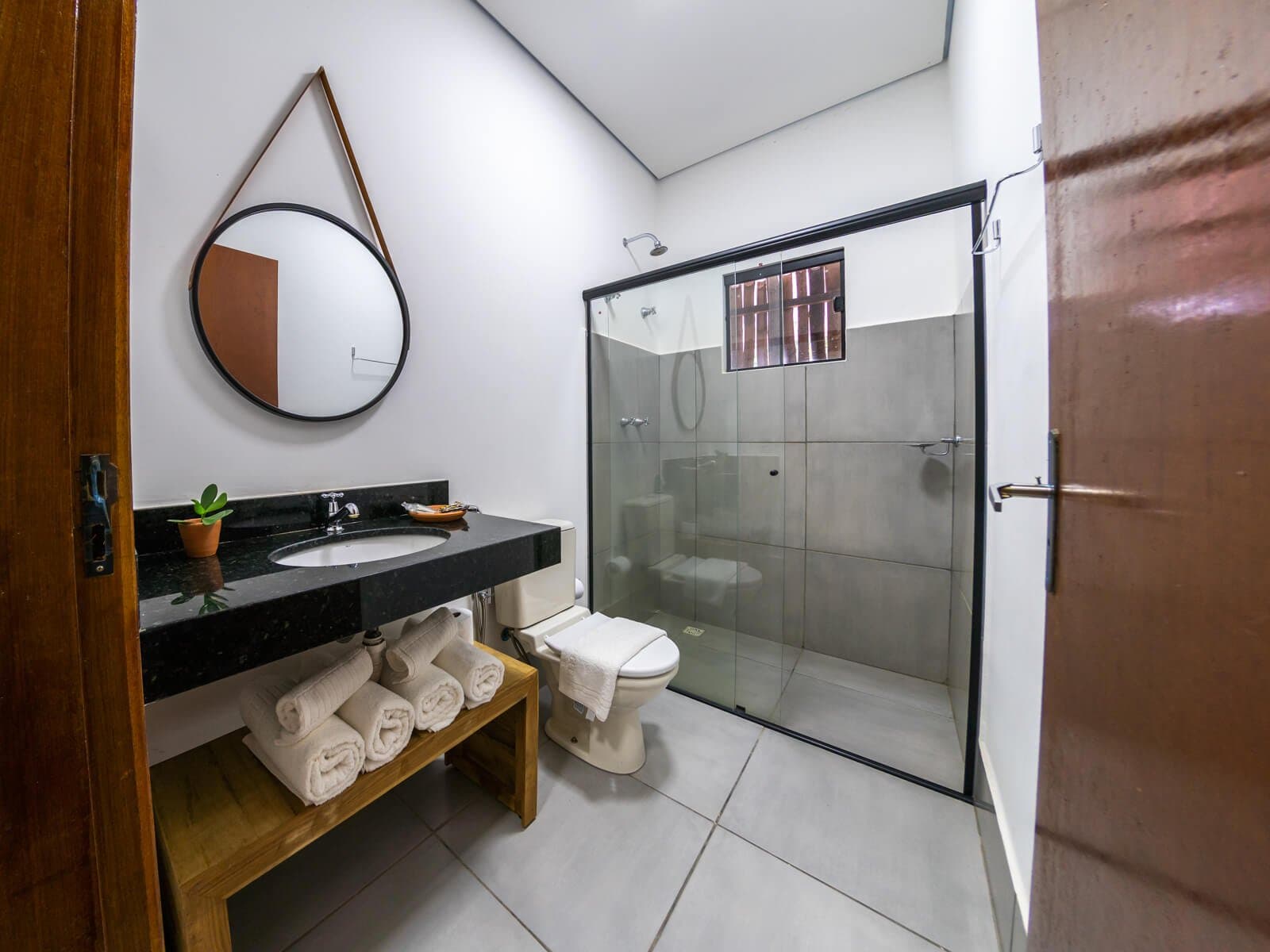 Fresh towels, a hot shower, and a small succulent keeping you company. Clean private bathroom at a Porto Jofre lodge on a pantanal jaguar safari, featuring a black granite vanity, round leather-strap mirror, glass-enclosed shower, and neatly rolled white towels on a wooden shelf