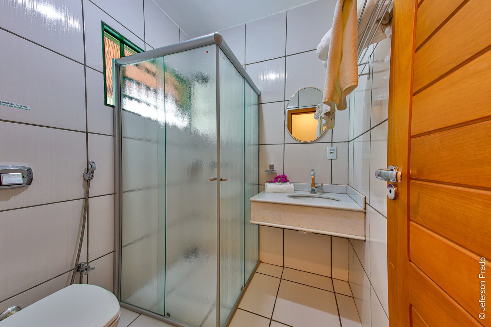Small but thoughtfully kept, so you come back from the trail ready to feel human again. Clean private en-suite bathroom at a Porto Jofre lodge, featuring a glass-enclosed shower cubicle, white tiled walls, marble vanity with a round mirror, and fresh towels on a heated rail