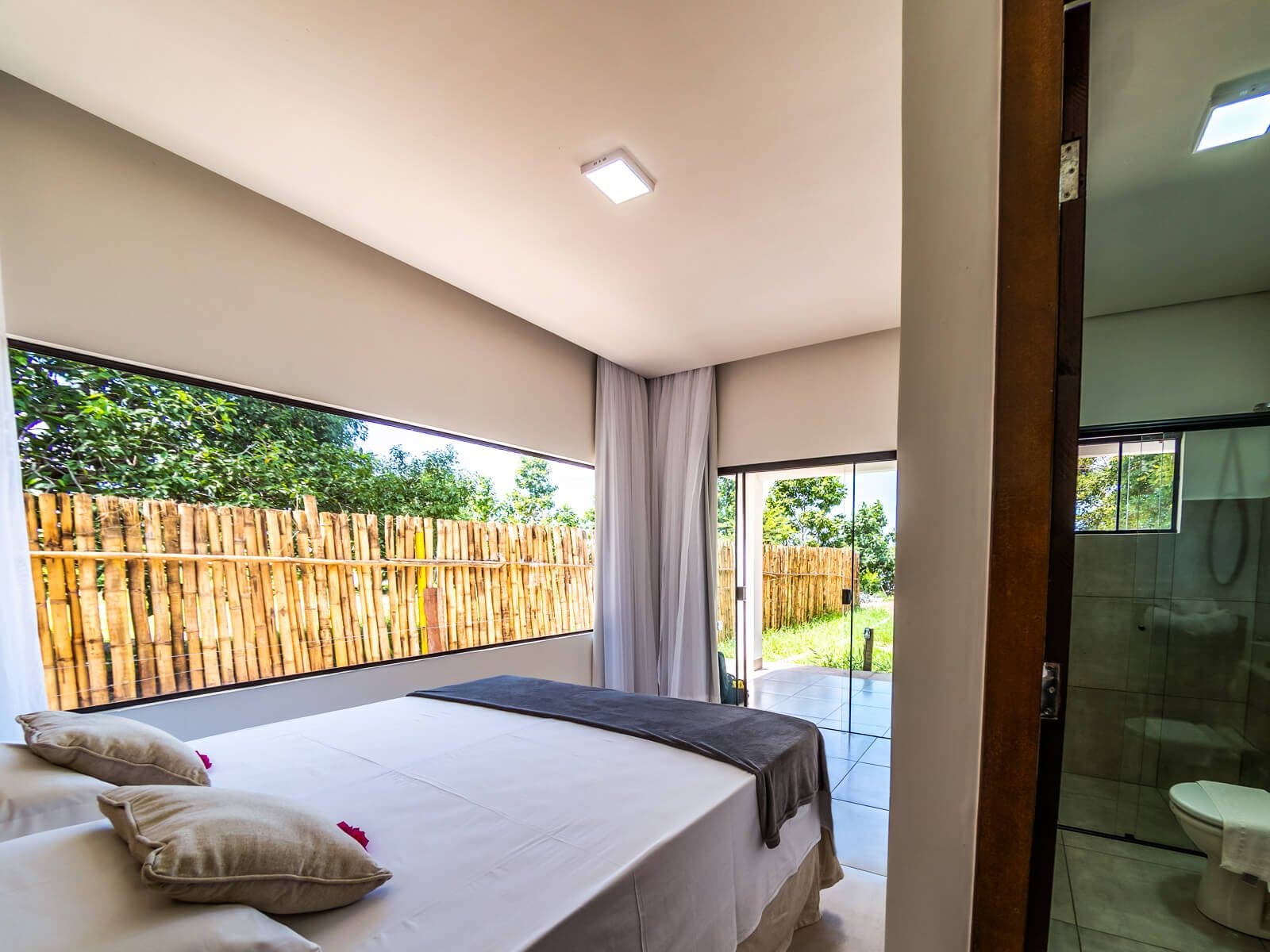 Clean lines, garden greenery, and a proper bed after a day in the field. A bright double bedroom at a pantanal jaguar safari porto jofre lodge, featuring white bedding, linen cushions, a wide picture window with a bamboo privacy screen, and a glass-walled en-suite bathroom