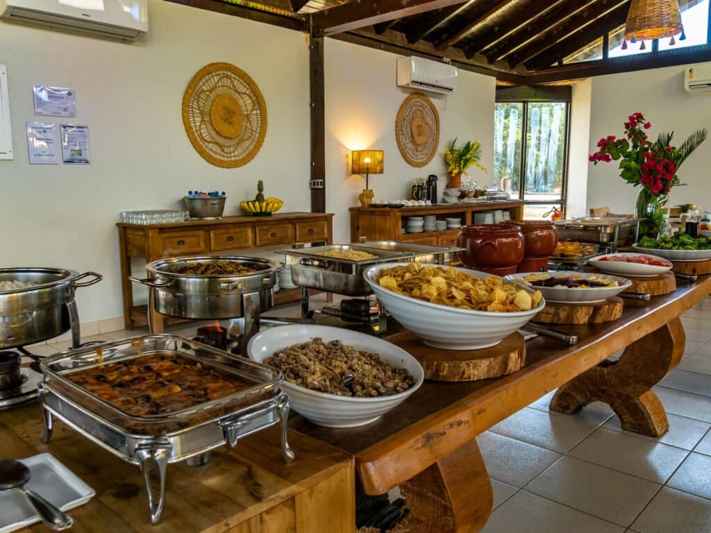 Fuel up properly before a full day of wildlife watching. A generous Brazilian buffet spread on a rustic wooden table at a pantanal jaguar safari porto jofre lodge, featuring chafing dishes, farofa, fried snacks, stewed dishes, and fresh flowers