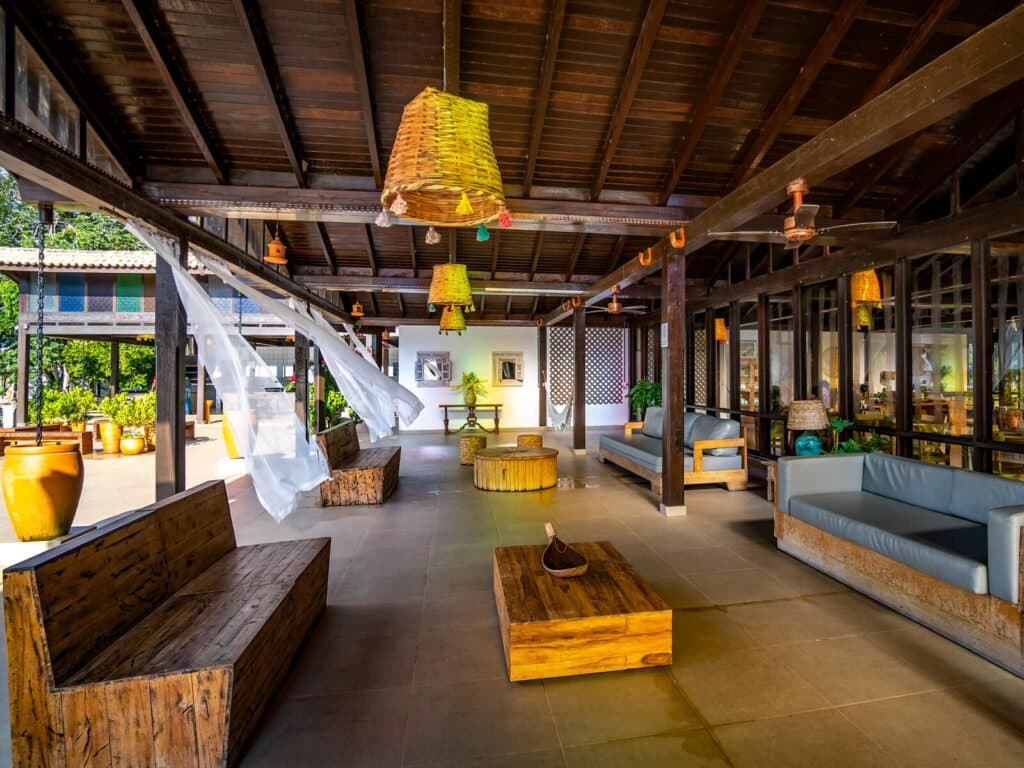 The kind of lobby that makes you forget you had a schedule. Open-air lodge lounge at a pantanal jaguar safari porto jofre property, featuring rustic hardwood benches, woven pendant lamps, and sheer curtains billowing in the breeze
