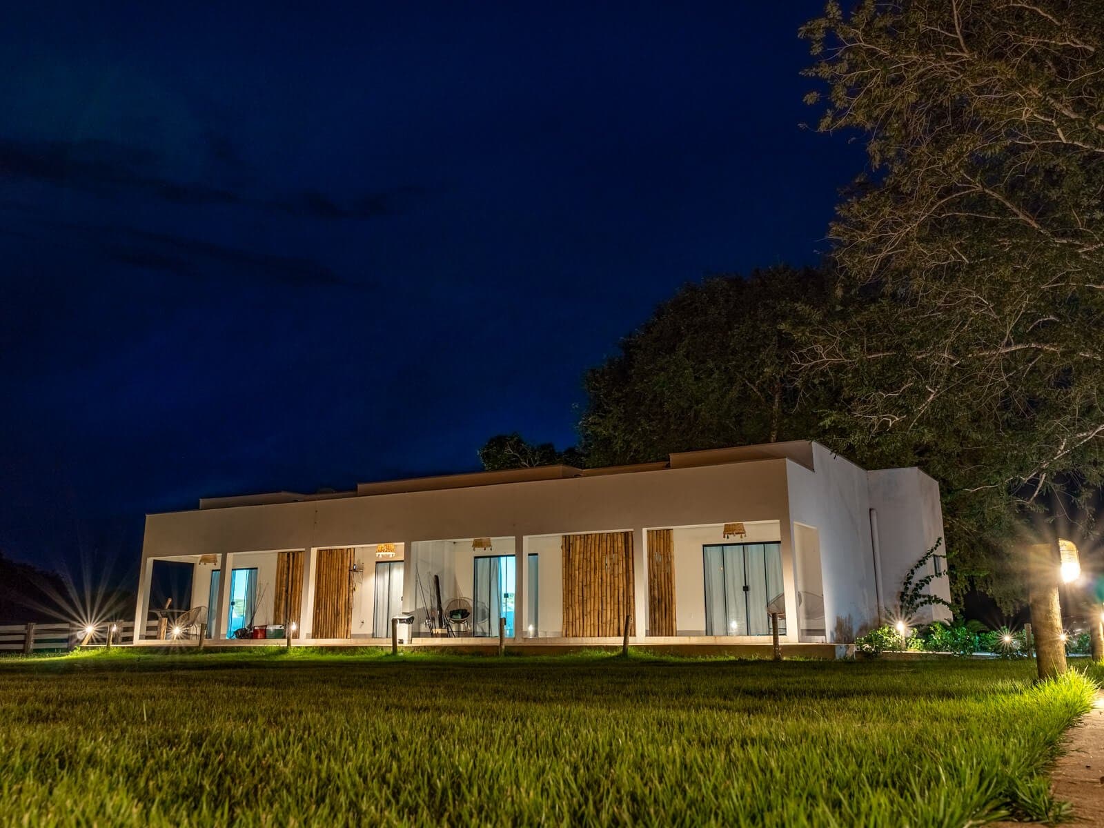 Nights out here are quiet, and your room is already waiting with the lights on. Illuminated white lodge with bamboo accents glowing under a deep blue night sky on a pantanal jaguar safari porto jofre, surrounded by green lawns and tall trees