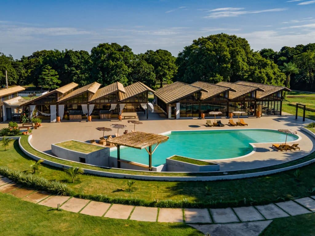 After a morning of wildlife, this pool earns every minute of your afternoon. Aerial view of a Pantanal jaguar safari Porto Jofre lodge featuring a curved turquoise pool, thatched-roof pavilions, sun loungers, and lush green lawns surrounded by native trees