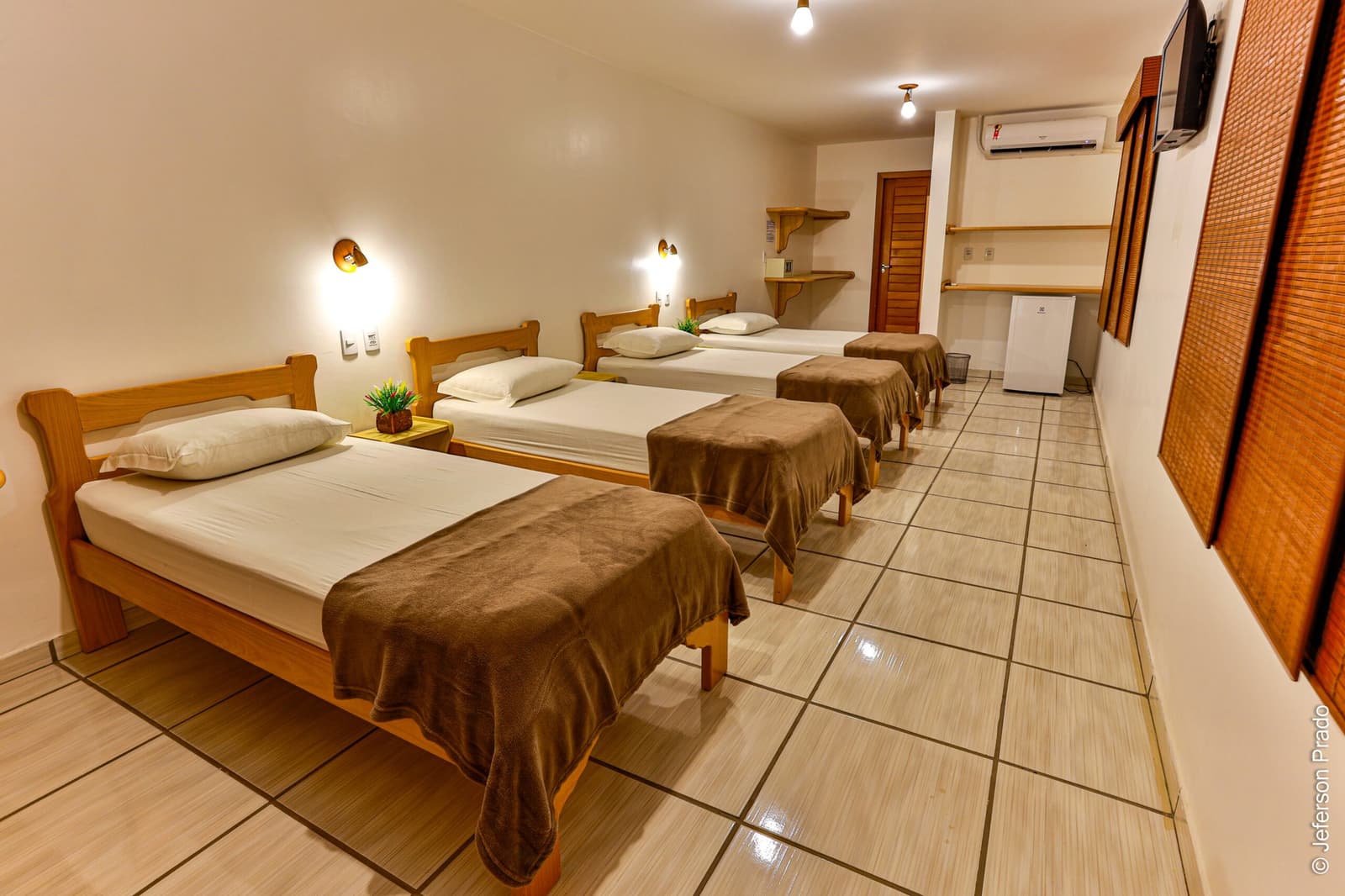 Four beds, one roof, and a full day of wildlife waiting outside. Spacious quad guestroom at a Porto Jofre lodge on a pantanal jaguar safari porto jofre trip, with four single wooden beds dressed in white linens and brown throws, wall-mounted reading lights, air conditioning, and a mini-fridge