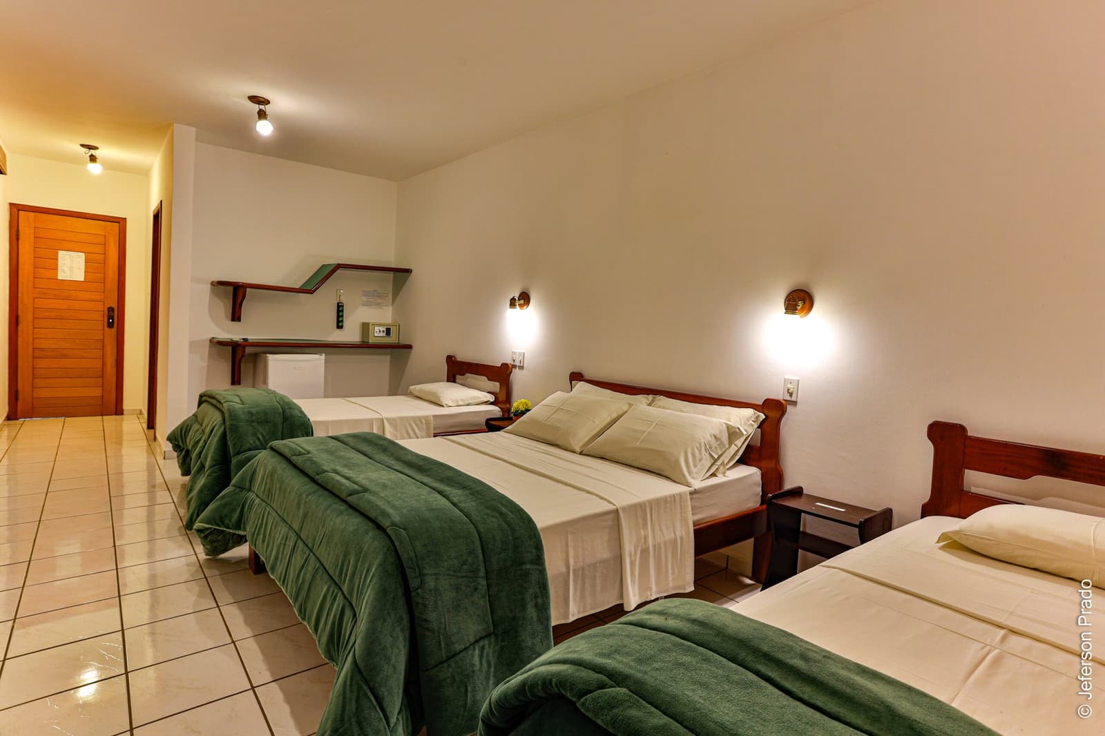 Clean, comfortable, and ready for early wake-up calls, the beds that earn their keep. Triple room at a Porto Jofre lodge on a pantanal jaguar safari porto jofre trip, with three single beds dressed in white linens and green blankets, wooden headboards, warm wall sconces, and a mini-fridge beneath a wall shelf