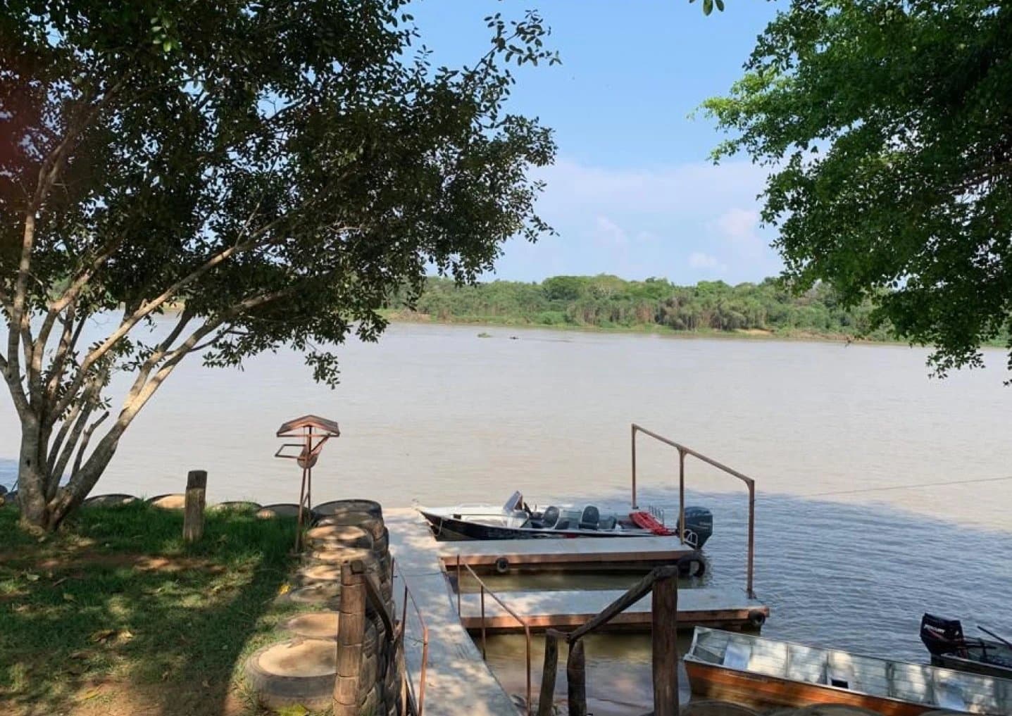 Your next adventure is already tied up and waiting. A wooden river dock with motorboats moored on a wide, brown-watered river, framed by riverside trees on a sunny day at the start of a pantanal jaguar safari porto jofre expedition