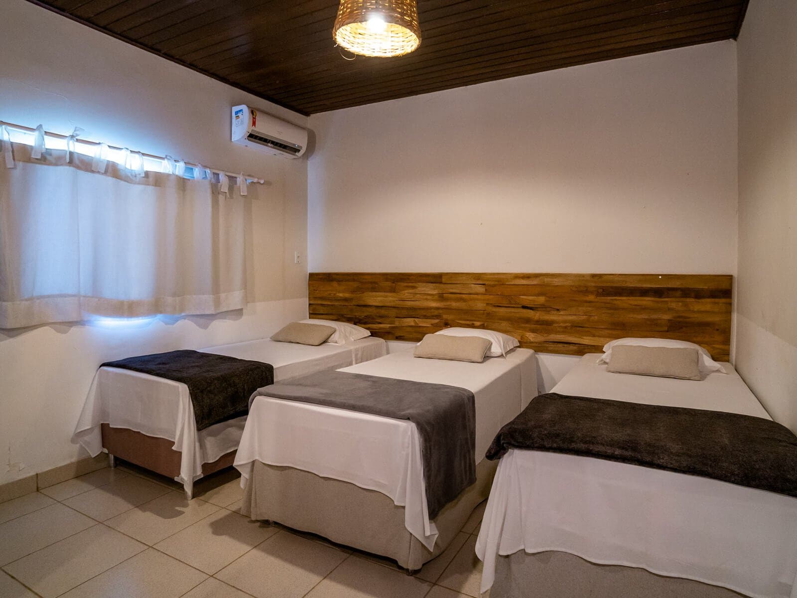 Three beds, one good night's sleep, the essentials, done right. A tidy triple room at a Porto Jofre lodge featuring three single beds with white linens, a rustic wood headboard, air conditioning, and a wicker pendant light — comfortable rest between pantanal jaguar safari days