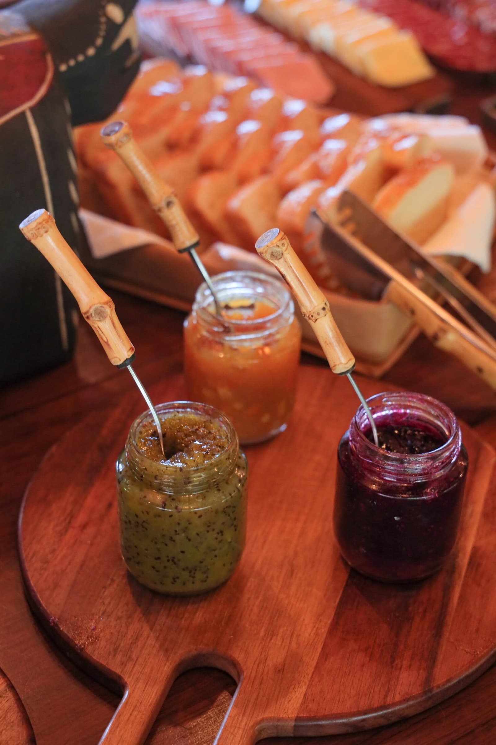 Three jams, one board, and the quiet pleasure of a slow morning. Three glass jars of homemade fruit preserves — green, orange, and berry — with bamboo-handled spoons on a round wooden board, served alongside sliced bread and cheese at a pantanal luxury safari lodge