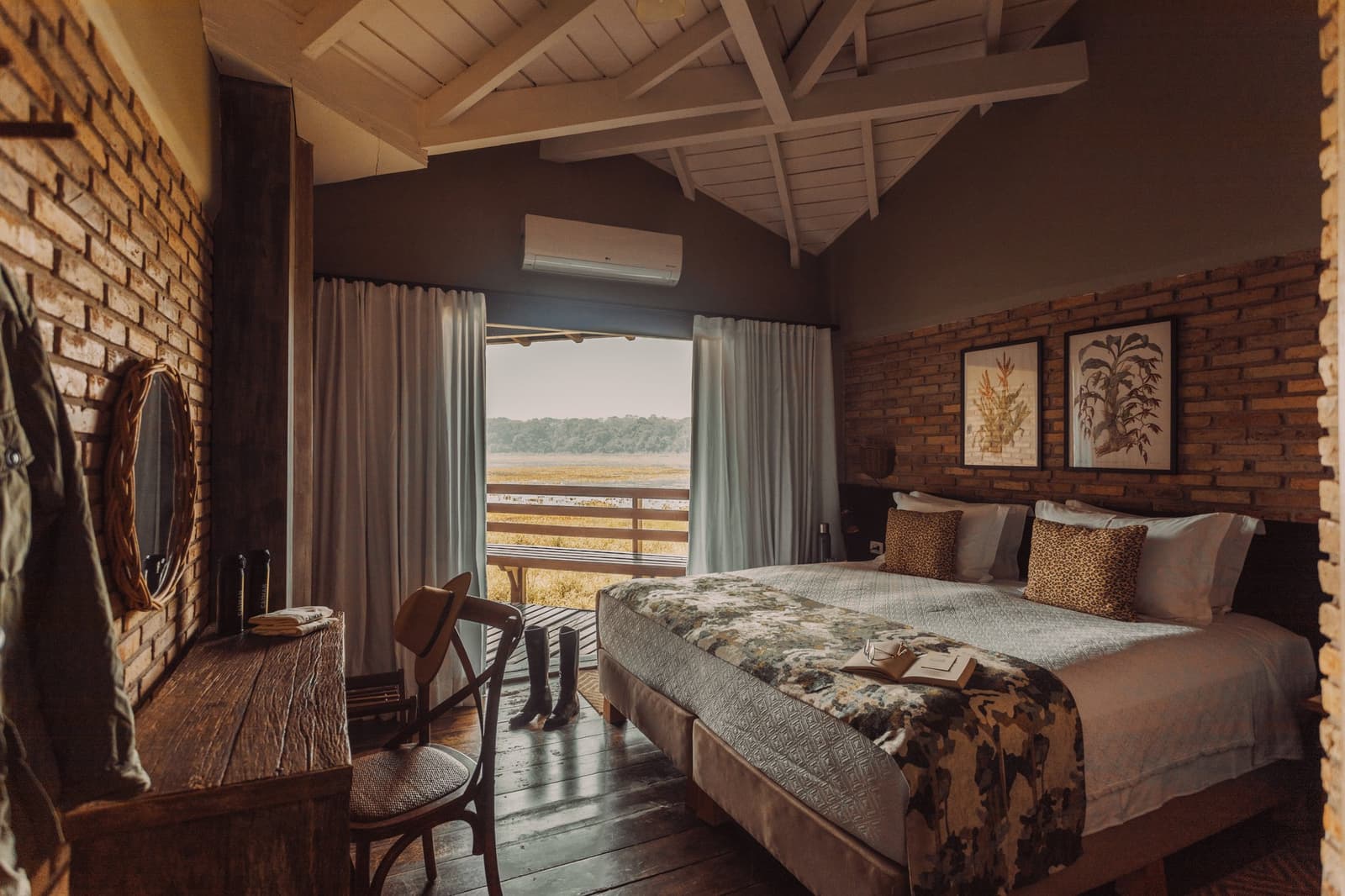 Rubber boots by the door, wetlands right outside, this is roughing it, Pantanal style. A rustic-chic lodge bedroom at Caiman featuring exposed brick walls, a king bed with leopard-print cushions, hardwood floors, and an open balcony door revealing a sweeping wetland view — the heart of a pantanal luxury safari experience