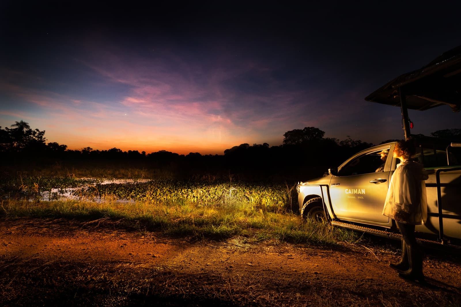 That quiet moment between the last light and the first sounds of the night. A guide stands beside a Caiman Ecological Refuge safari vehicle at dusk, with a fiery orange and violet sky reflected over a wetland lagoon covered in aquatic vegetation during a pantanal luxury safari
