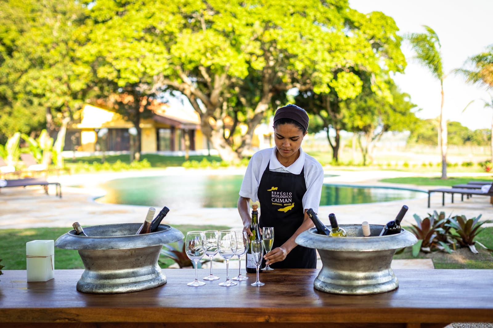 The welcome drink is cold, the afternoon is warm, and someone already thought of everything. A Caiman Ecological Refuge staff member in a branded apron arranging sparkling wine glasses and chilled bottles in metal ice buckets on a wooden bar, with a swimming pool and lush grounds visible behind her — part of the pantanal luxury safari experience