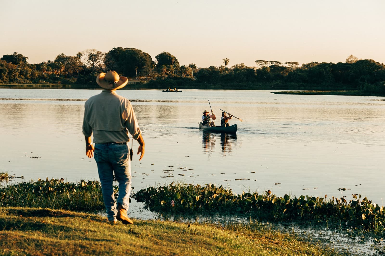The canoe goes first. The guide already knows what's out there. A guide in a wide-brimmed hat watches from the bank as two guests paddle a canoe across a calm wetland lagoon during a pantanal luxury safari at golden hour