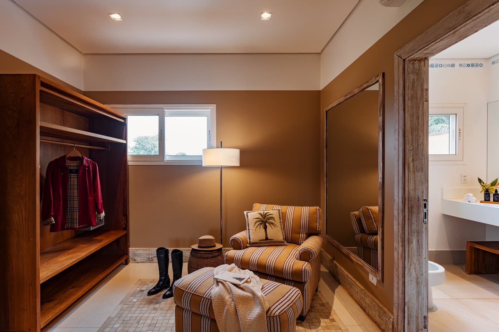 Pack light, dress well, your wardrobe here has better stories than most. A warmly lit dressing area in a pantanal luxury safari lodge, featuring an open timber wardrobe, a striped armchair with a palm-motif cushion, riding boots, a straw hat, and an ensuite bathroom glimpsed through a rustic wooden doorframe