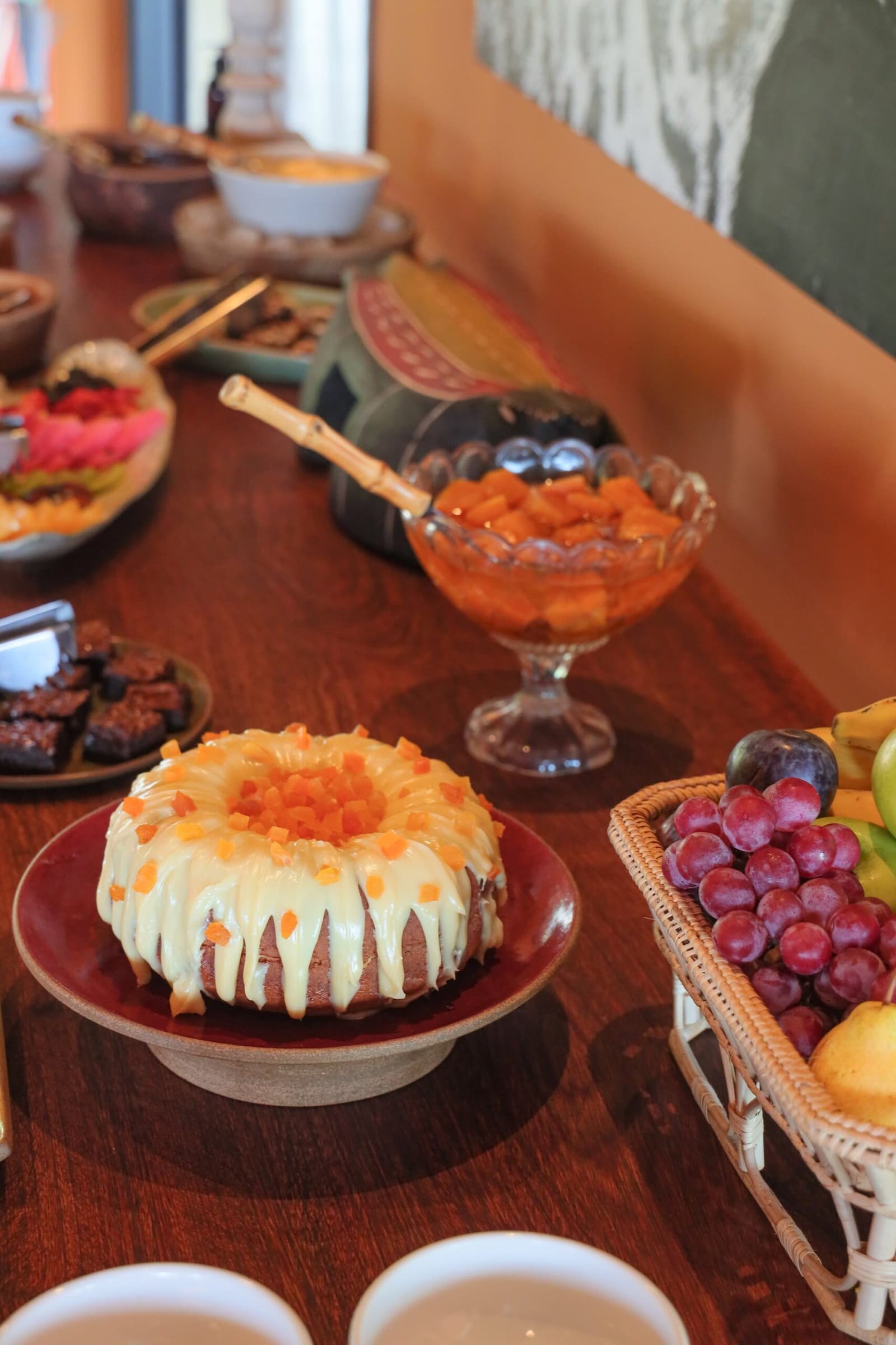 Mornings here start with a spread that makes it hard to head out on safari. A glazed bundt cake topped with diced papaya sits on a red ceramic stand at a pantanal luxury safari lodge breakfast buffet, surrounded by fresh grapes, diced fruit, and brownies
